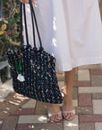 Person holding a black floral-patterned tote bag on a paved walkway.