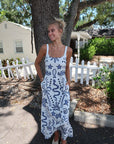 Woman in a white dress with blue patterns standing outdoors near a tree and house.