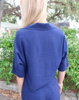 Person wearing a blue suit standing outdoors with greenery in the background, back view