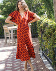 A woman modeling a short-sleeve V-neck midi dress with a bold orange and dark red abstract pattern, standing on a brick patio with green hedges in the background.