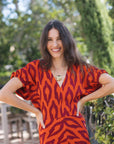 A woman wearing a short-sleeve V-neck dress with an orange and dark red abstract pattern, standing outdoors with her hands on her hips against a blurred green background.