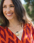 A smiling woman with long dark hair wearing a textured orange and burgundy patterned top and a gold pendant necklace, photographed outdoors with a blurred green background.