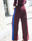 A person wearing wide-leg maroon pants with a bold blue side stripe, paired with a matching maroon top and beige heels, standing outdoors on a paved walkway.