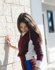 A woman wearing a burgundy and white color-blocked sweatshirt and matching sweatpants with a blue side stripe, leaning against a light stone wall in bright sunlight.