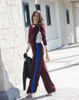 model wearing burgundy pants with a blue stripe holding a leather jacket and wearing a burgundy sweater with white details with a shopping center as the background