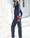 A woman wearing a navy blue long-sleeve top and matching wide-leg pants, holding a textured burgundy clutch bag, standing outdoors next to a light-colored stone wall.