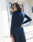A woman wearing a navy blue mock neck long-sleeve top and dark blue wide-leg jeans, holding a pink patterned clutch bag, photographed outdoors against a bright white building.