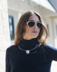 A woman with brown hair wearing aviator sunglasses with a silver frame and a silver circular pendant necklace, dressed in a black turtleneck sweater, photographed outdoors against a blurred light-colored building background.