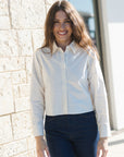 A woman with long brown hair smiling, wearing a white long-sleeve button-down shirt and dark blue jeans, standing outdoors against a light beige stone wall.