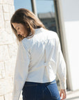 A woman wearing a white long-sleeve button-up shirt and dark blue jeans, photographed from behind in an outdoor setting with a light stone wall and window in the background.
