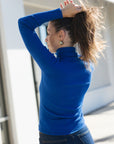 A woman wearing a bright royal blue ribbed turtleneck sweater and dark jeans, seen from the back and side as she adjusts her hair. She is wearing small gold hoop earrings.