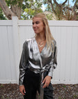 Woman wearing a silver metallic top and black pants standing outdoors with a white fence and trees in the background.