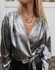 Person wearing a silver metallic top with layered necklaces against a white fence background, close up