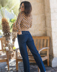 A woman wearing a beige and brown geometric patterned crop top and dark blue straight-leg jeans, standing on a tiled patio next to a wooden bench and stone wall.