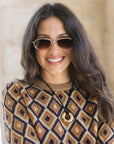A woman with long brown hair wearing a brown and beige geometric patterned knit sweater, gold-framed sunglasses, and a gold pendant necklace, smiling against a soft beige background.