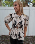 Woman wearing a patterned blouse standing outdoors with a white fence and trees in the background