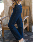 A person wearing dark blue straight-leg jeans, a brown and beige diamond-patterned knit sweater, and brown slippers, standing next to a wooden bench in an outdoor patio setting.