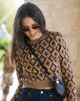 A woman wearing a brown, beige, and black geometric diamond-patterned knit sweater, dark blue jeans, and sunglasses, carrying a small snakeskin-patterned crossbody bag.