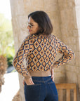 A woman wearing a cropped long-sleeve knit sweater with a geometric diamond pattern in beige, brown, and navy, paired with dark blue jeans, photographed outdoors against a stone pillar.