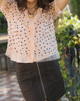 A woman wearing a sheer light peach short-sleeve blouse with black polka dots and a black asymmetrical mini skirt, standing outdoors with her arms raised.