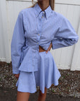 Person wearing a blue striped shirt and skirt set against a white fence.