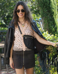 A woman wearing a black leather jacket, a beige polka-dot blouse, a black mini skirt, and a black woven crossbody bag, standing outdoors in front of green foliage.