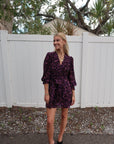 Woman wearing a purple floral dress standing in front of a white fence with trees in the background, full body