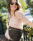A woman outdoors wearing a brown woven crossbody bag with gold hardware over a light peach polka-dot blouse.