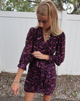 Woman wearing a purple patterned dress standing outdoors with a white fence and trees in the background. sleeve detail