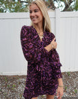 Woman wearing a purple patterned dress outdoors with a white fence and trees in the background.
