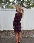 Woman wearing a purple dress with a pattern, standing in front of a white fence. full body back