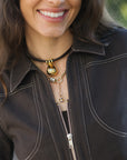 A close-up lifestyle shot of a woman wearing layered necklaces, including a black choker with a gold teardrop pendant and a gold beaded chain, paired with a dark brown jacket.