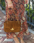 A person outdoors holding a brown suede shoulder bag with a gold chain strap and a gold triangular front accent. The person is wearing a long rust-red patterned maxi skirt and clear high-heeled sandals.