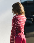 A woman shown from behind wearing a fuzzy long-sleeve knit sweater with thin white horizontal stripes on a deep red base, paired with bright pink pants, standing outdoors against a white wall.