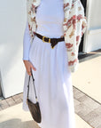 Person wearing a white dress with a floral cardigan and black belt, holding a brown bag.