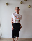 Person wearing a white t-shirt with colorful designs and a black skirt in a minimal indoor setting.