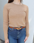 Person wearing a beige long-sleeve top and blue jeans with a belt, standing against a light-colored wall.