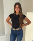 Woman wearing a black top and blue jeans with a gold belt standing against a neutral wall.