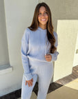 Woman wearing a light blue ribbed sweater and pants standing outdoors.