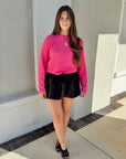 Woman wearing a pink sweater and black shorts standing against a light-colored wall.