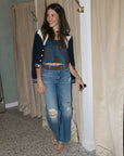 A woman stands in a fitting room wearing a navy and white cardigan over a blue patterned crop top, paired with distressed blue jeans and tan heeled sandals.