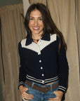 A woman smiling while wearing a navy blue and white knit varsity-style cardigan with white buttons, paired with blue jeans and a brown belt, standing in front of beige curtains.
