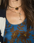 A close-up of a model wearing layered gold necklaces, including a choker with a shell pendant and a delicate chain with a T-bar and small round charm, over a blue floral top.