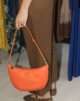 A person wearing brown patterned trousers and leopard-print flats holds a bright orange crescent-shaped leather shoulder bag with a matching strap in a clothing store.