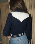 A woman viewed from behind wearing a navy blue ribbed knit cardigan with a large white chevron pattern on the back, paired with blue jeans.