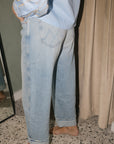 A person wearing light-wash wide-leg denim jeans with rolled cuffs, paired with a light blue embellished top, standing barefoot in an indoor setting with a mirror and beige curtain visible.