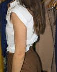 A woman wearing a white sleeveless collared blouse with a ruffled hem and brown polka-dot trousers, standing in a clothing store next to racks of garments.