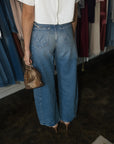 A person shown from the back wearing wide-leg blue jeans, a white short-sleeve top, and nude heels, while holding a small metallic gold handbag in a clothing store.