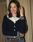 A woman smiling and posing in a navy blue and white knit cardigan with striped ribbed trim, paired with blue jeans and a brown belt, set against a background of beige curtains.