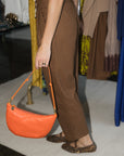 A person wearing brown textured wide-leg pants and leopard-print flats, holding a bright orange crescent-shaped leather shoulder bag with a gold-tone ring clasp.